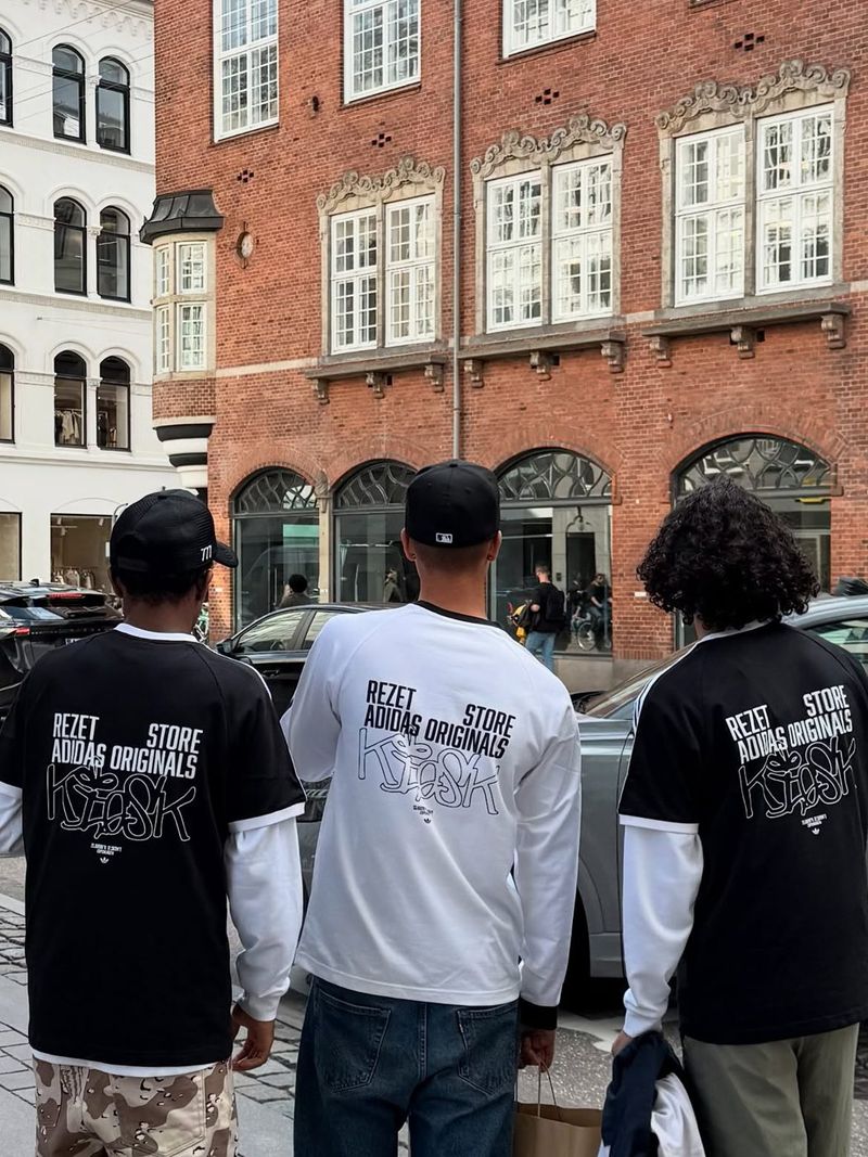 Three people seen from behind on a city street, wearing Rezet Store × adidas Originals Kiosk T-shirts.