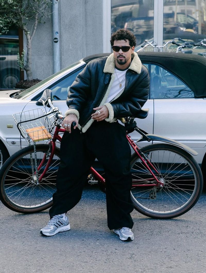 A man in a shearling-collared leather jacket sits on a red bicycle on a city street, wearing Adistar Control 5 sneakers.
