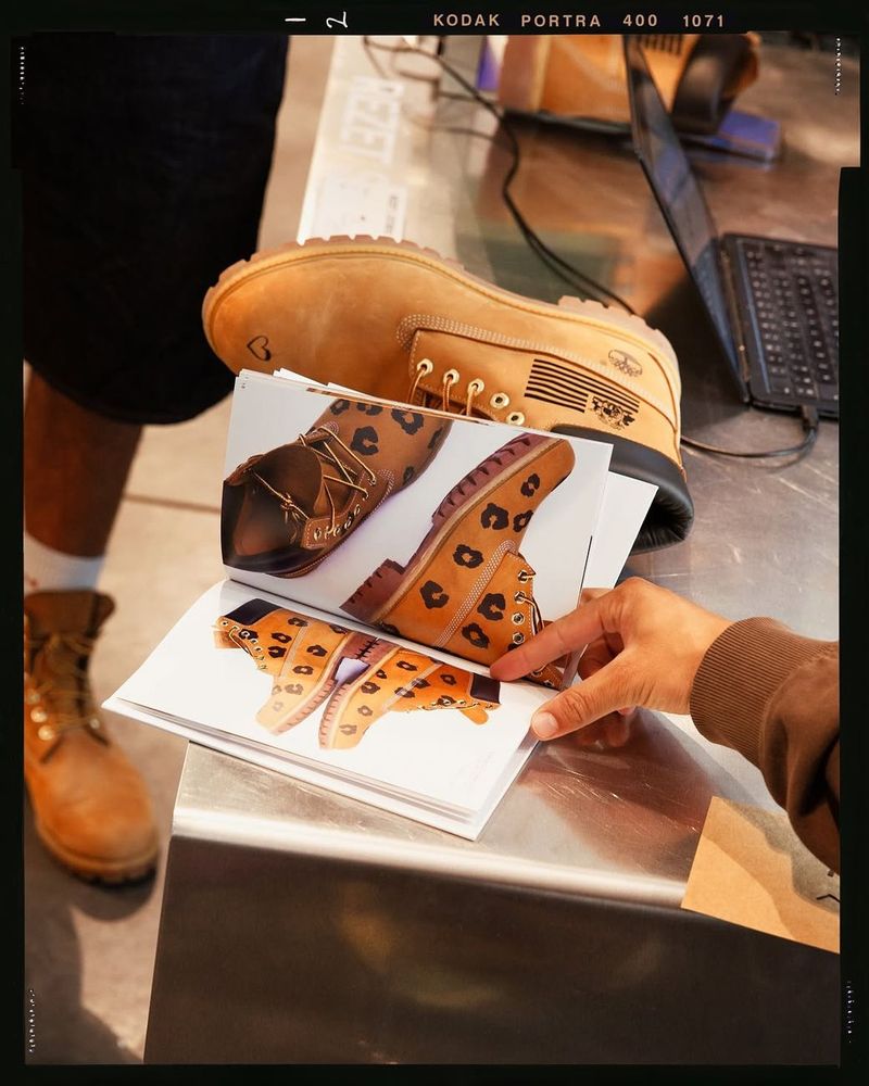 A hand holds printed pages showing customized boots, next to a pair of wheat Timberland boots on a steel counter.